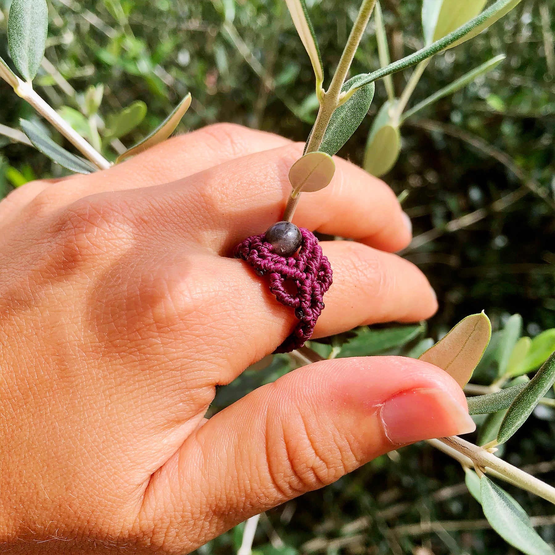 Anello macramé artigianale in marrone scuro con dettaglio di pietra su una mano tra foglie di ulivo