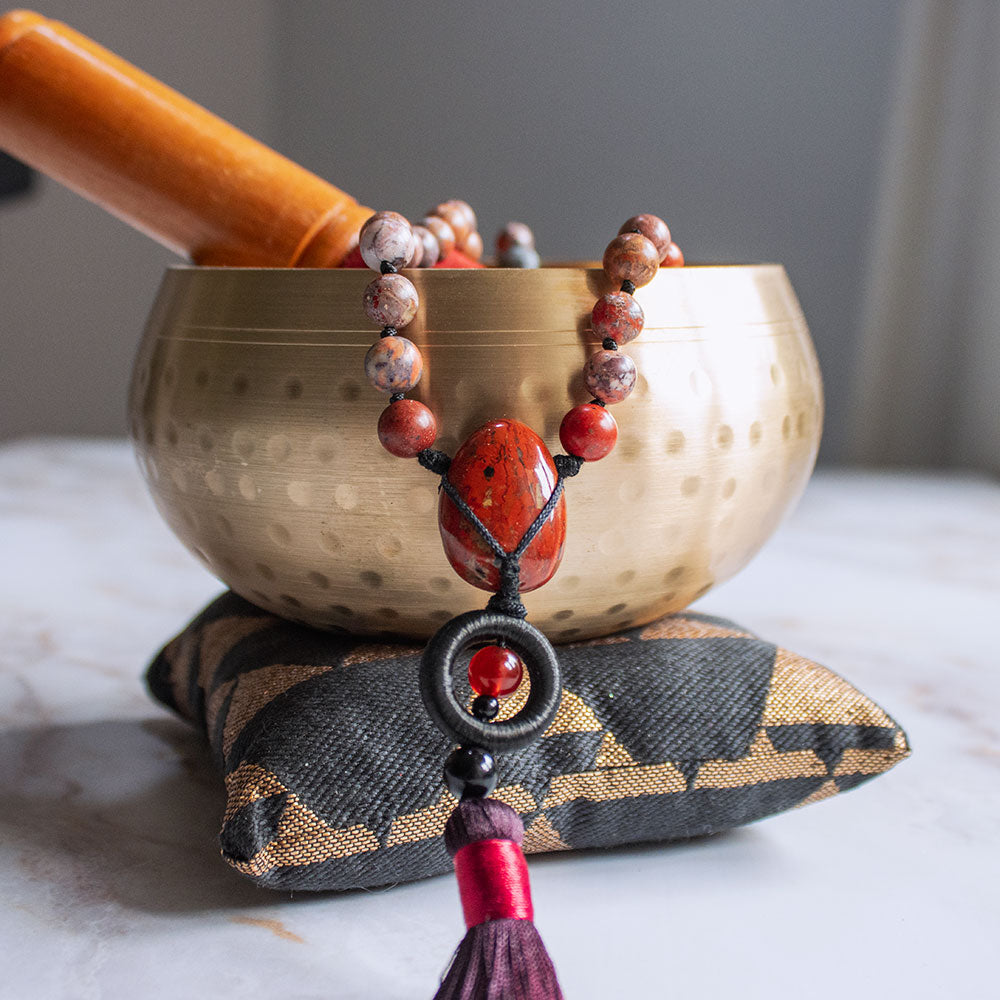 Collana mala diaspro rosso e ossidiana con perline su un cuscino decorativo accanto a una ciotola tibetana