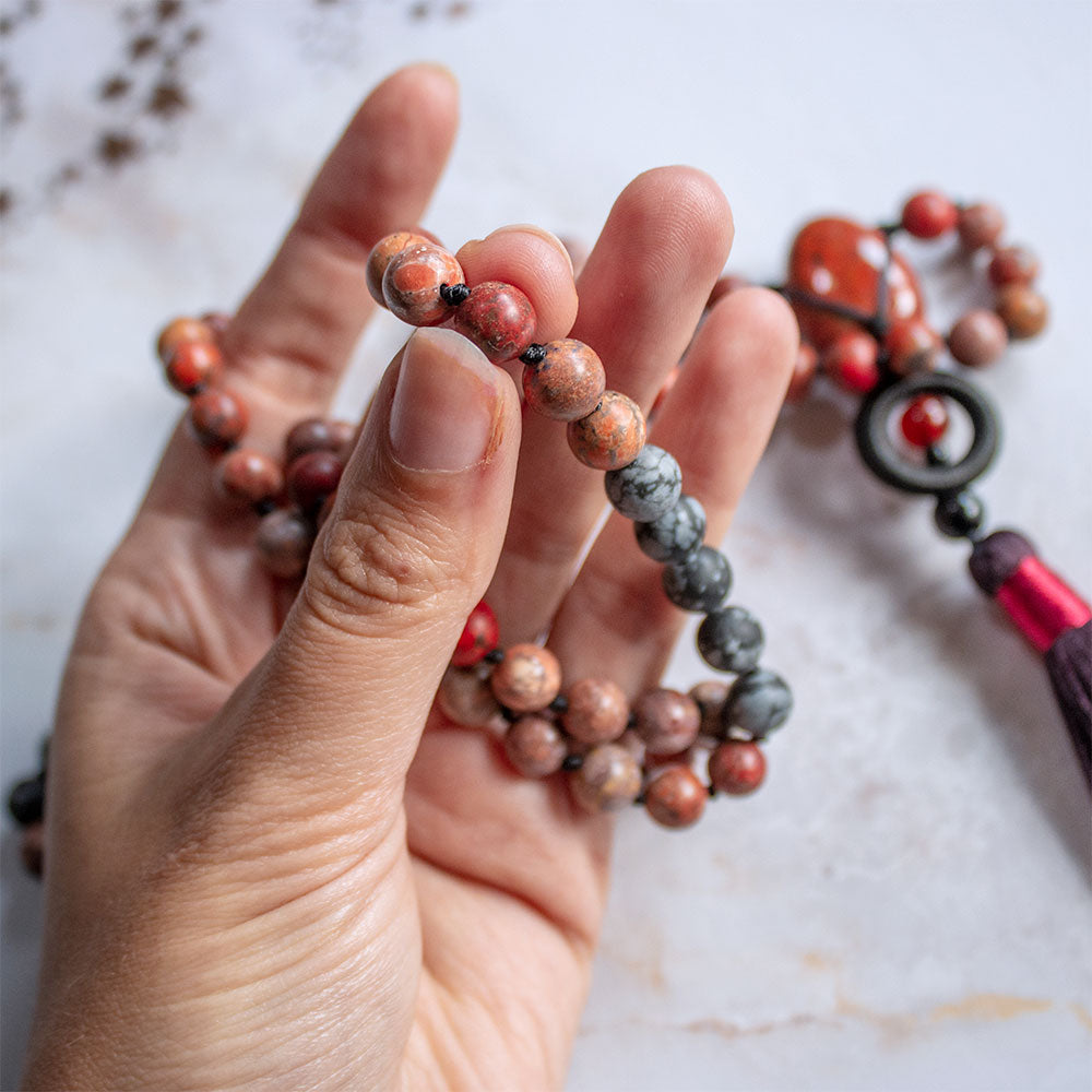 Collana mala diaspro rosso e ossidiana con perline colorate in una mano su sfondo marmorizzato
